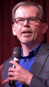 Medium Closeup photo of Jason on stage wearing dark glasses, a blue shirt and dark blue suit.